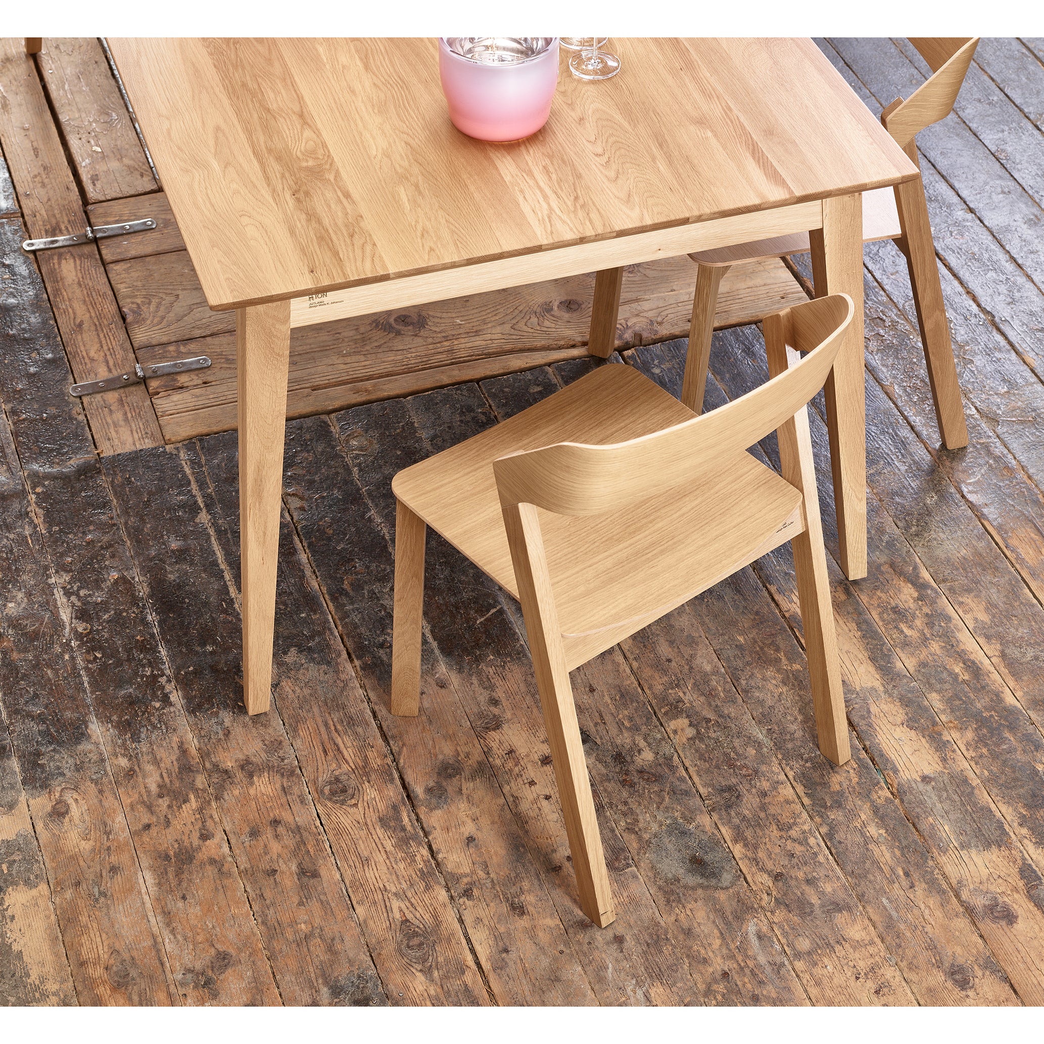 The Jylland Table by TON, with its light wooden finish, exudes Scandinavian elegance on a rustic floor. Positioned at an angle, the extendable table is adorned with a pink candle in a glass container and two glasses, enhancing the serene setting.