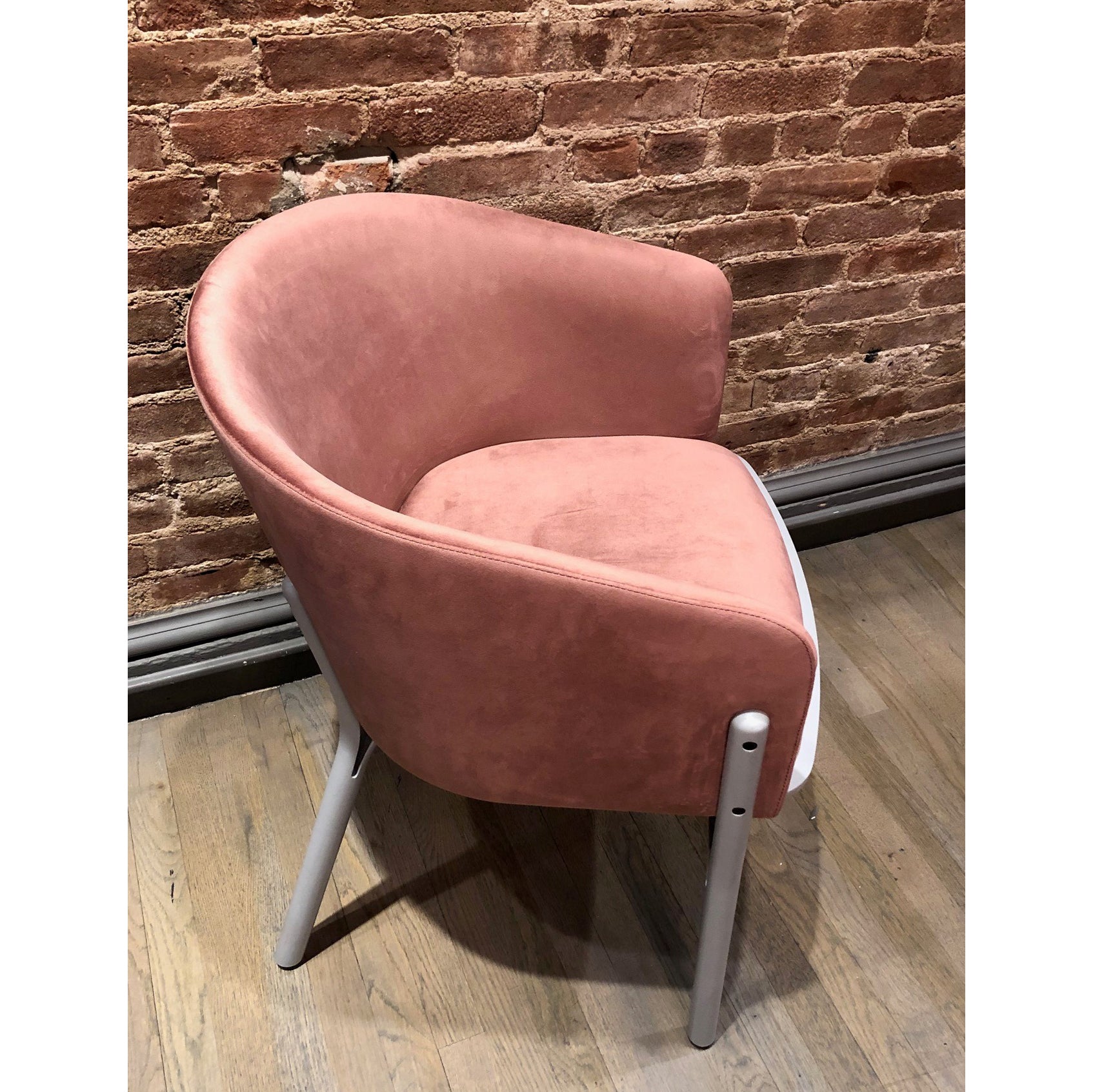The pink Armchair Split from TON, featuring a velvet upholstery and white metal legs, sits gracefully on a wooden floor against a rustic brick wall, merging modern design with classic charm.
