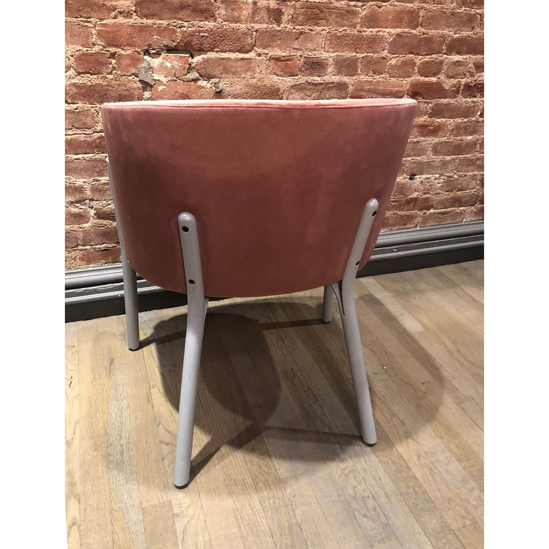 A pink velvet Armchair Split from TON, featuring light gray legs crafted with manual wood bending techniques, sits on a wooden floor against an exposed brick wall. The chair is viewed from the back.