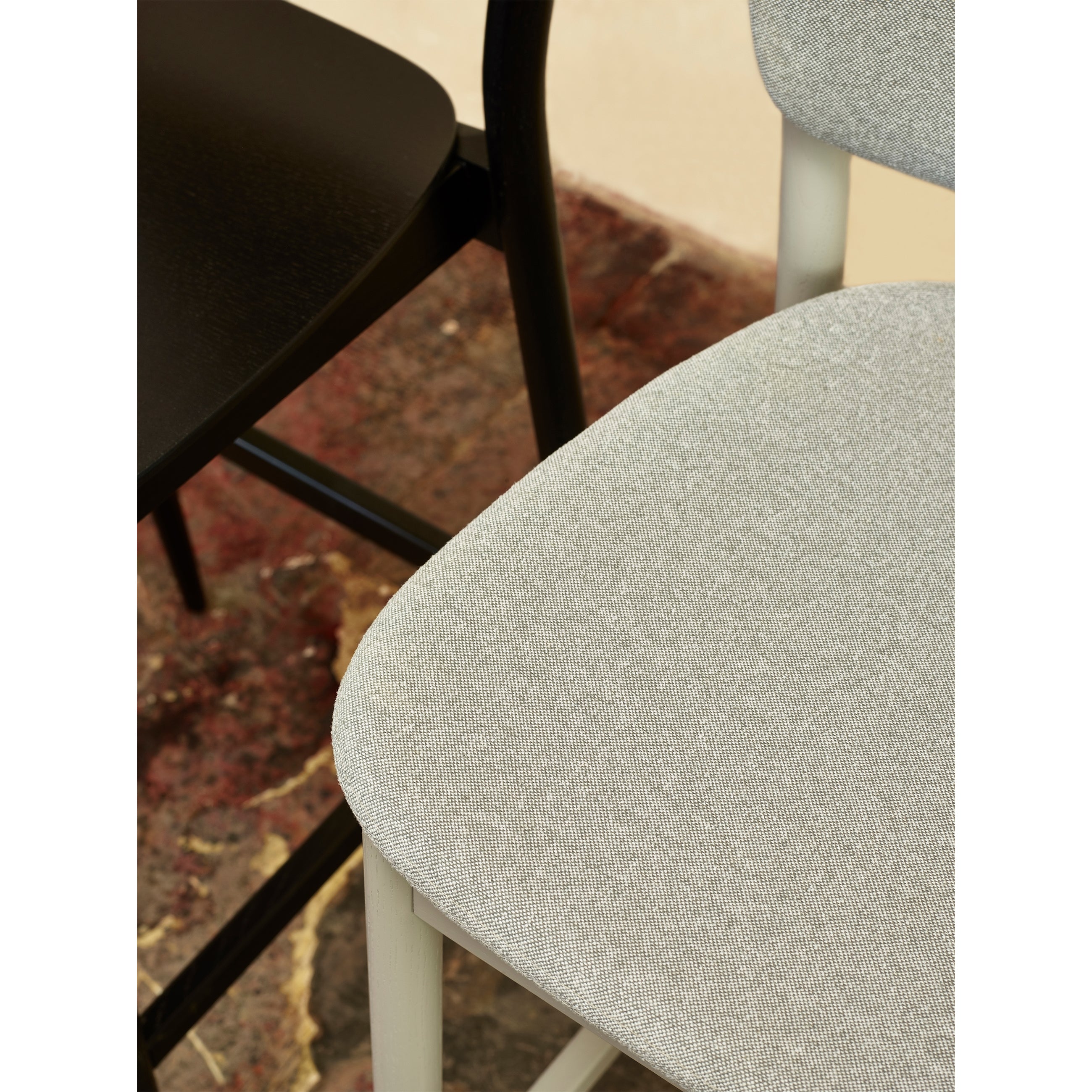 A close-up shows two chairs: one has a dark wooden seat, and the other features a light gray upholstered seat, both on a textured floor. This contrast in materials and colors echoes the elegant design of the Barstool Valencia by TON.