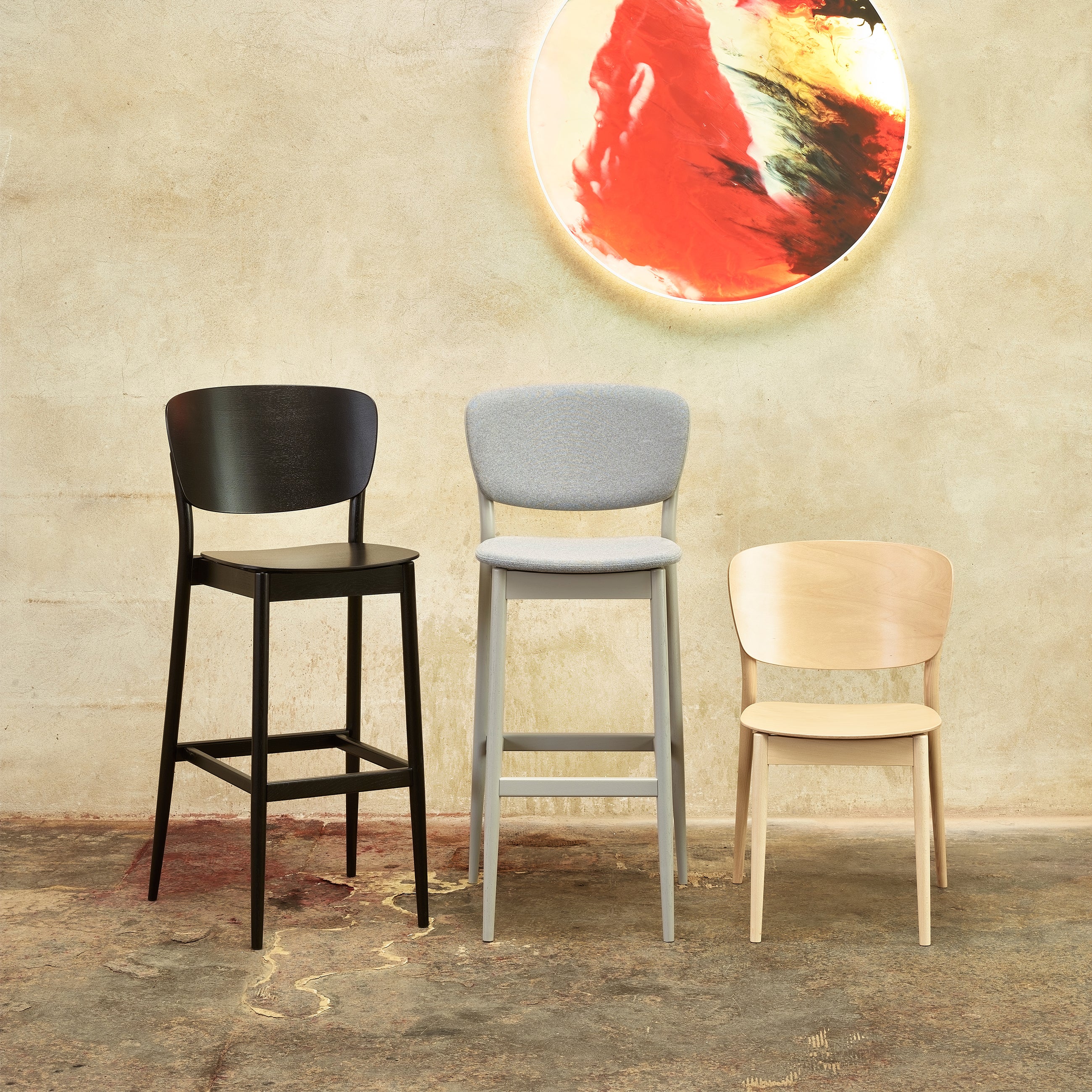 Against a textured wall sit three distinct chairs: the TON Brands sleek Barstool Valencia on the left, a medium-height grey chair in the center, and a small light wood chair on the right. Above them glows an abstract art piece reminiscent of Yonoh designer creations.