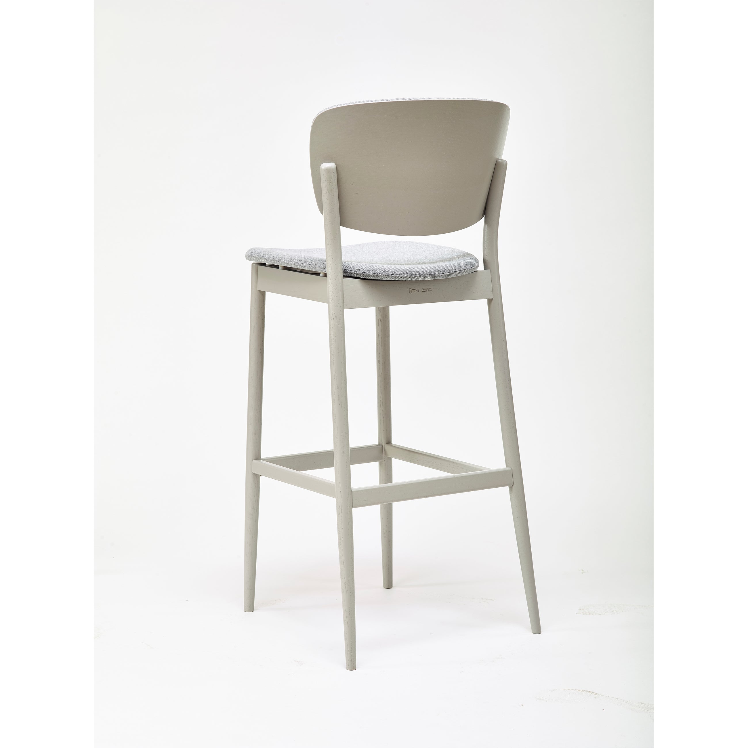 The Barstool Valencia from TON is a modern, sleek high barstool with a light gray frame and cushioned seat. It features minimalist design elements, including a curved backrest and straight legs, against a white background, designed by the talented Yonoh.