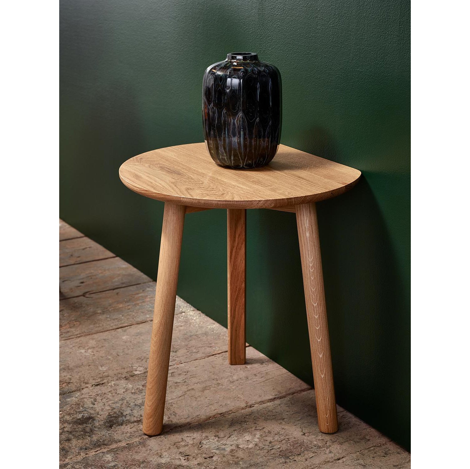The TON YYY Coffee Table, with its small round wooden design and three legs, stands against a dark green wall. A glossy black vase with a textured pattern sits on top, while the natural oak flooring enhances the spaces rustic charm and warmth.
