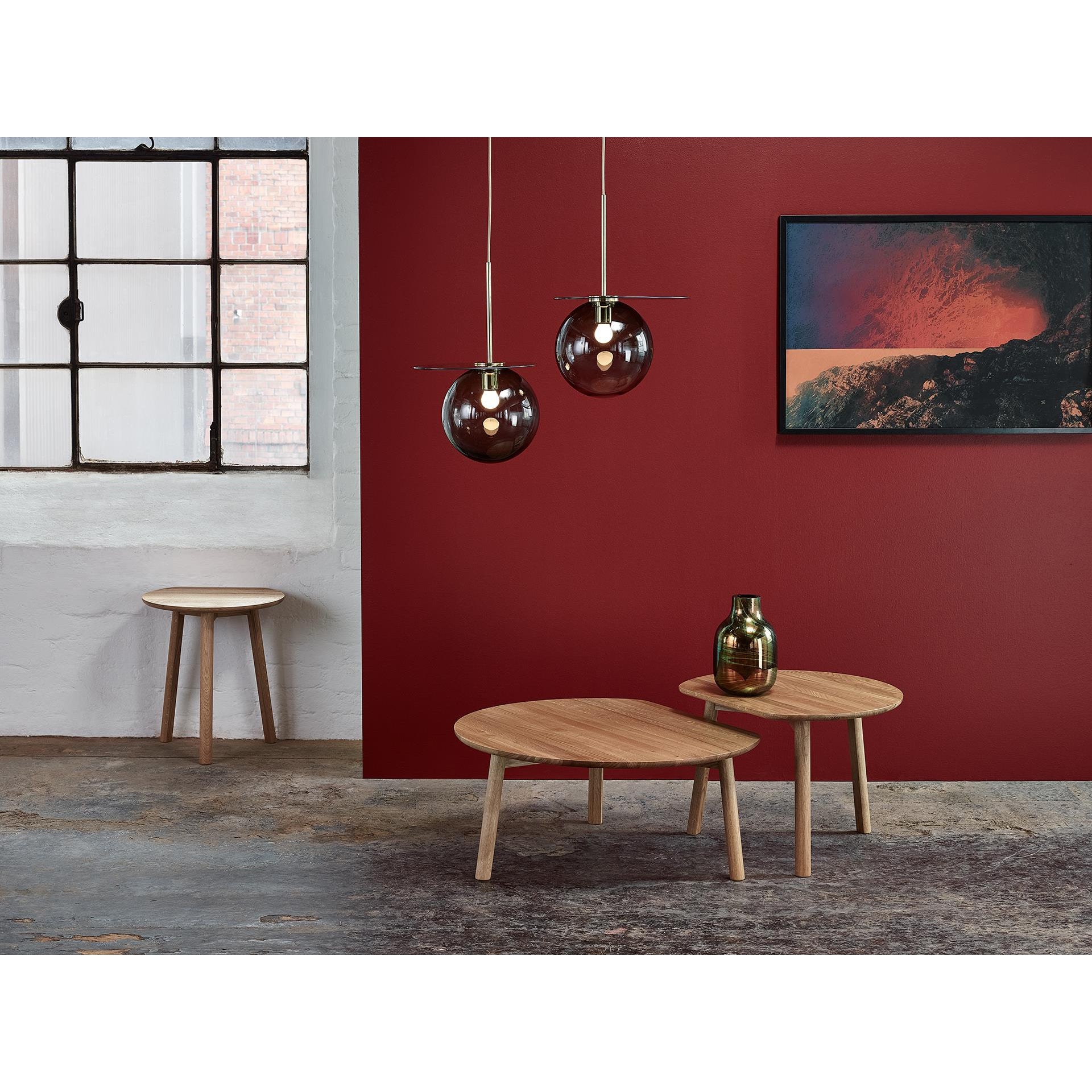 A minimalist room with red walls showcases two YYY Coffee Tables by TON in natural oak at different heights. Spherical pendant lights hang overhead, a modern painting is wall-mounted, and a small round beech side table sits by the window.