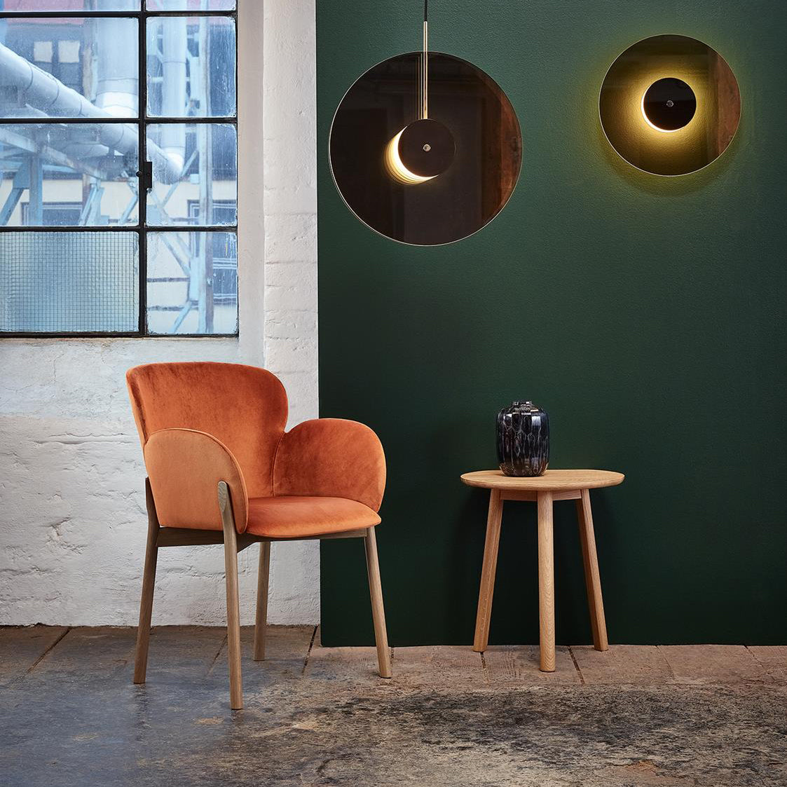 A cozy setting with a natural oak upholstered chair, a TON YYY Coffee Table, and a black vase. The wall is dark green and features two circular wall lights. A metal-framed window on the left illuminates the room.