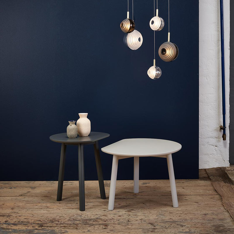 A modern gray YYY Coffee Table and a white one by TON stand against a dark blue wall, each holding small vases. Above them, pendant lights with metallic and white finishes illuminate the rustic natural oak floor.