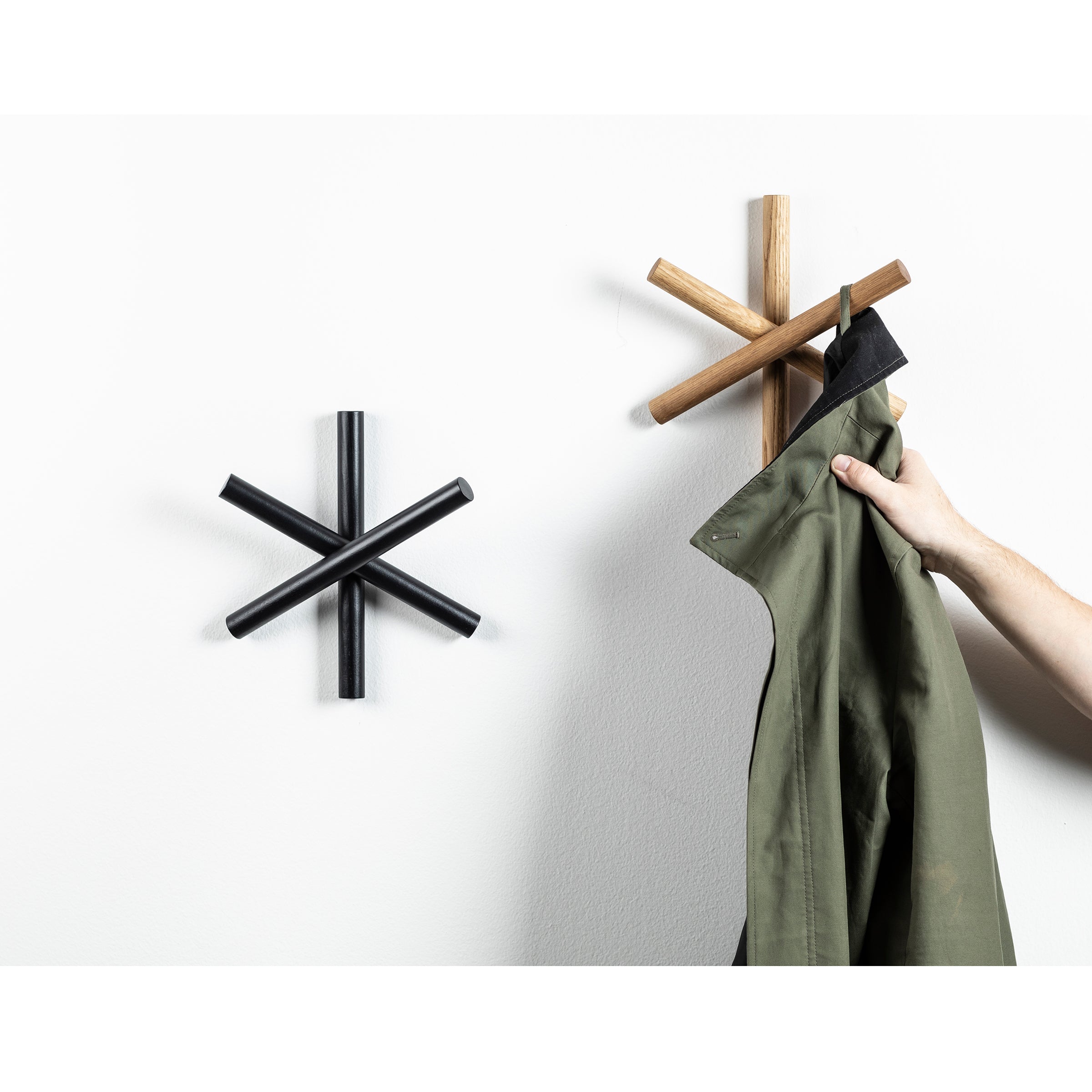 A person hangs a green jacket on a star-shaped wooden coat hook from TON, crafted using quality wood offcuts. An identical black Logs hook awaits its first use. Resembling stylish asterisk symbols, both hooks add charm to any HelpSoul workspace.
