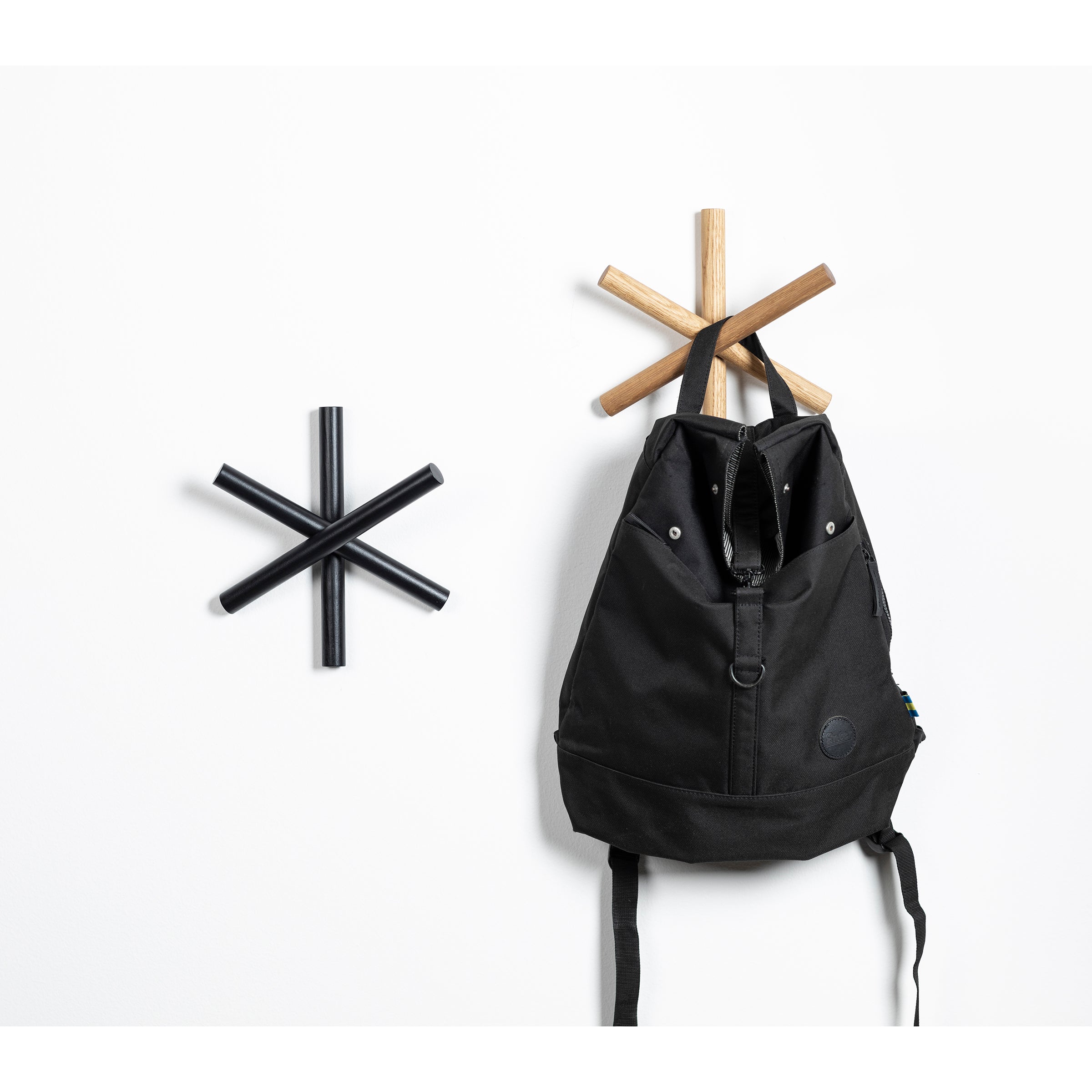 In the HelpSoul workspace, a black backpack hangs on TONs Logs wooden coat rack, uniquely shaped like a six-pointed star and crafted from quality wood offcuts. Another identical rack is mounted empty beside it on the white wall.