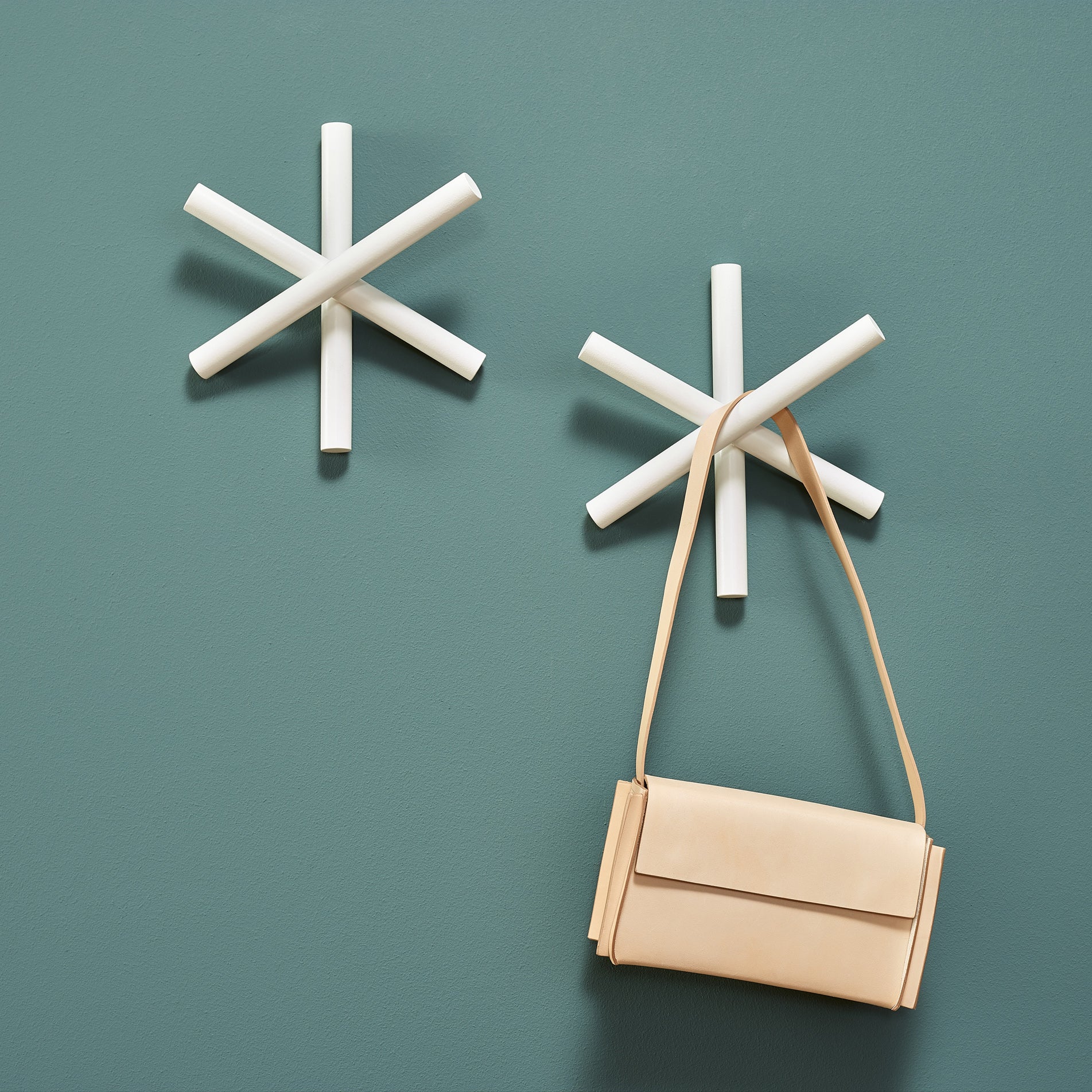 Two white, star-shaped wall hooks from TONs Logs collection adorn a green wall. Made of quality wood offcuts, they offer a modern minimalist design. A beige handbag hangs from the lower hook, blending sustainability with style.