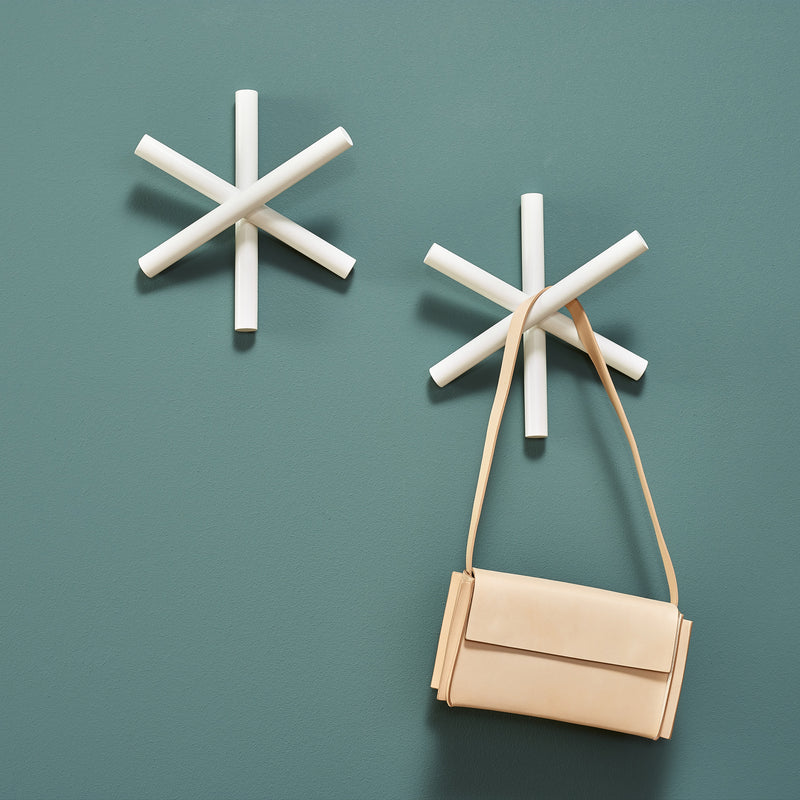 Two white, star-shaped wall hooks from TONs Logs collection adorn a green wall. Made of quality wood offcuts, they offer a modern minimalist design. A beige handbag hangs from the lower hook, blending sustainability with style.