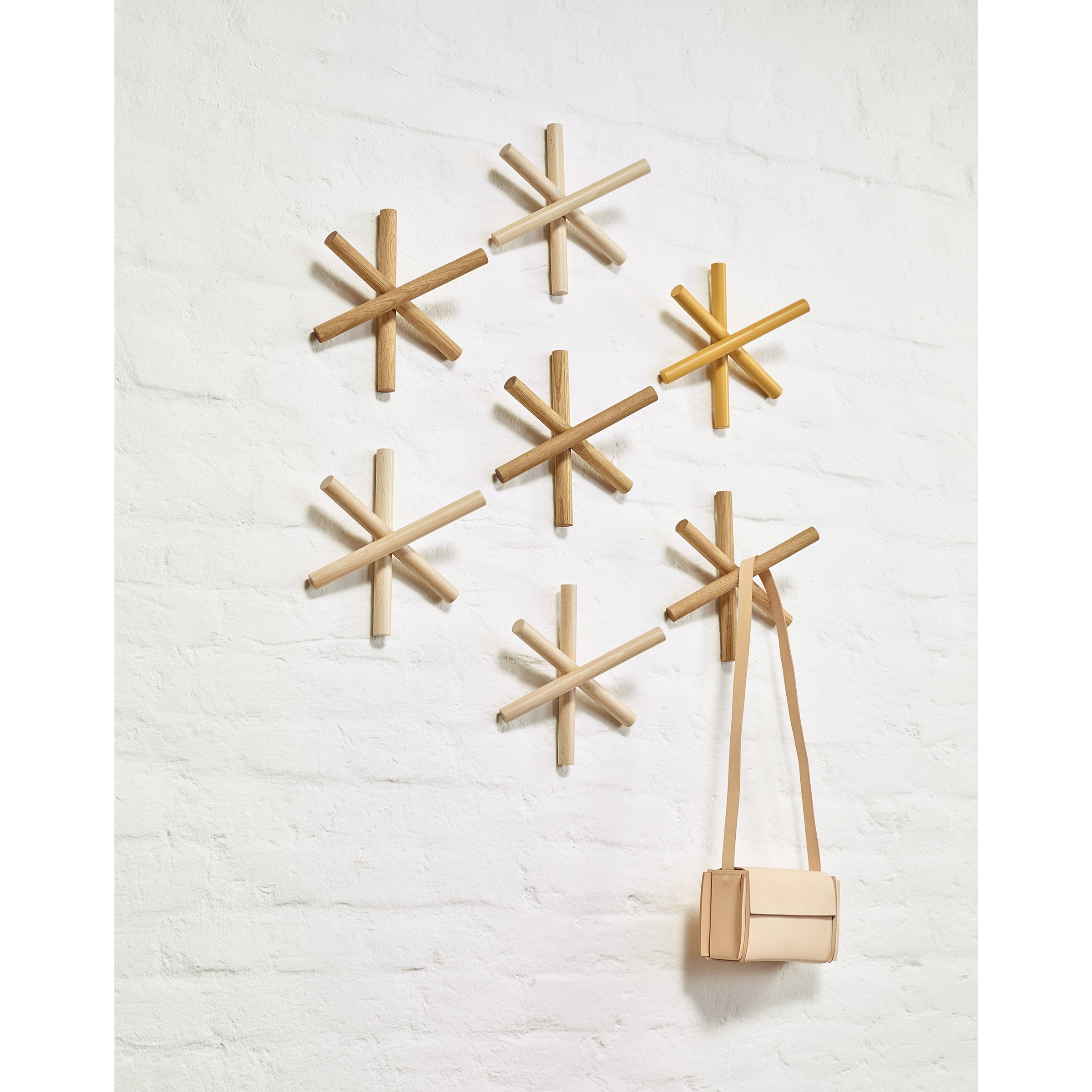 The TON Logs, innovative wooden coat hooks made from quality wood offcuts, create a geometric star pattern on a white brick wall. A beige handbag hangs gracefully from one hook, enhancing the HelpSoul workspaces charm.