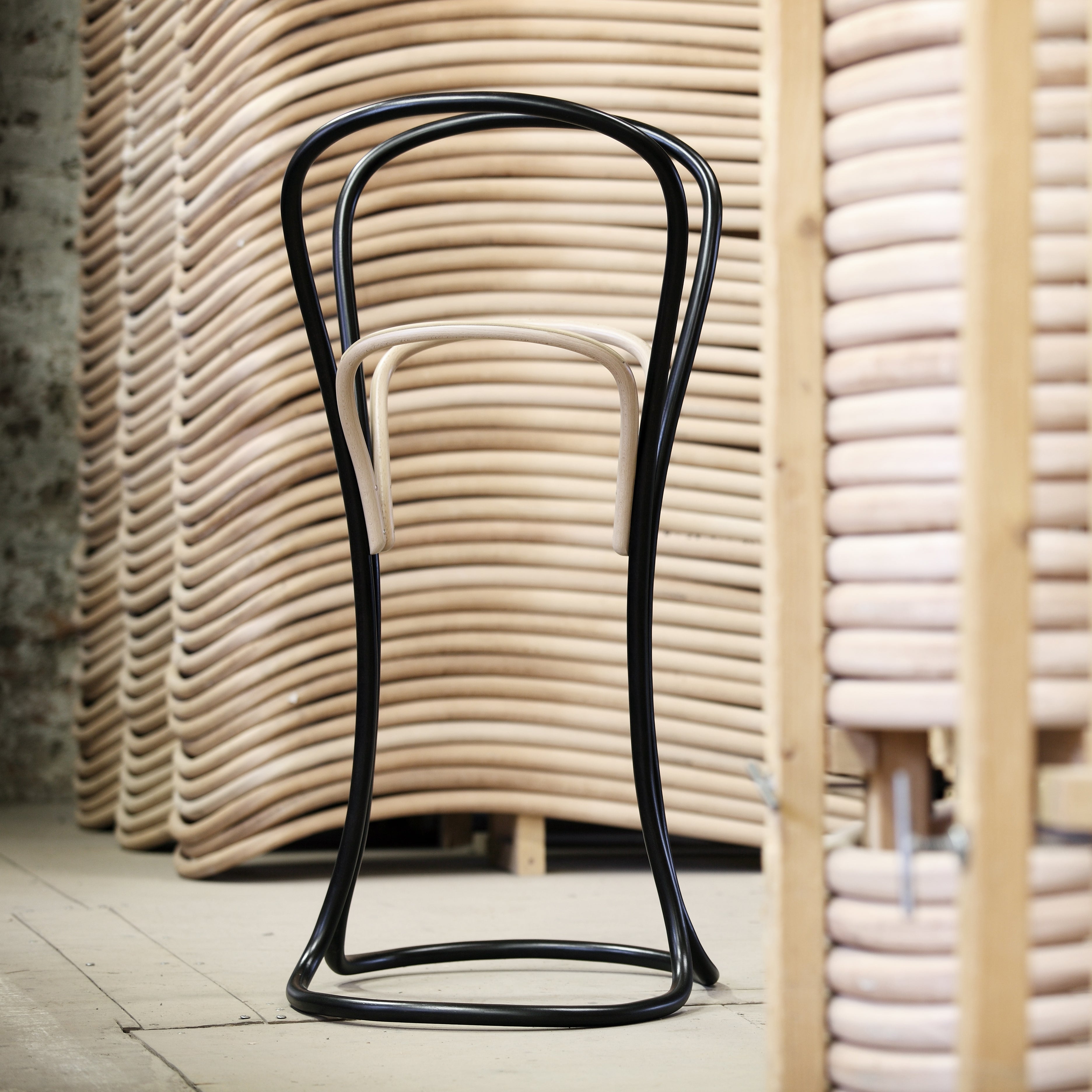 The Petalo, a distinctive bar stool by TON, features a black curvy frame and light wood seat, crafted with traditional wood bending techniques, displayed in front of neatly stacked wooden chair frames in a workshop.