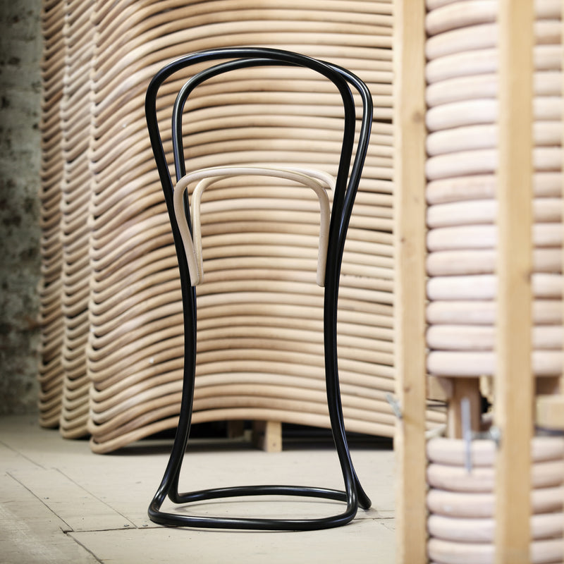 The Petalo, a distinctive bar stool by TON, features a black curvy frame and light wood seat, crafted with traditional wood bending techniques, displayed in front of neatly stacked wooden chair frames in a workshop.