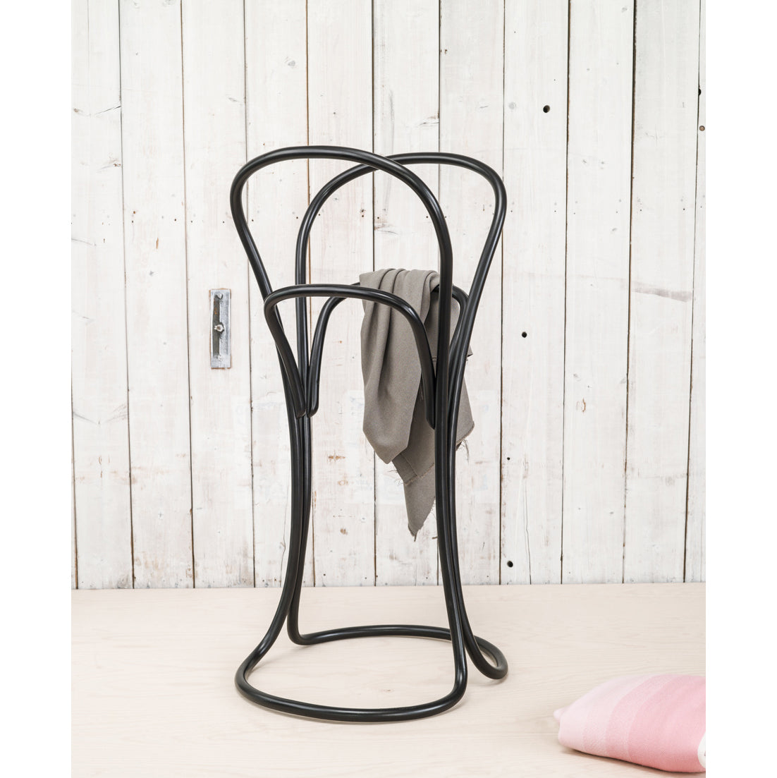 The Petalo by TON is a sleek black metal valet stand with a minimalist curved design, holding a gray garment and resting on a light wooden floor against rustic white planks. A folded pink fabric peeks out below.