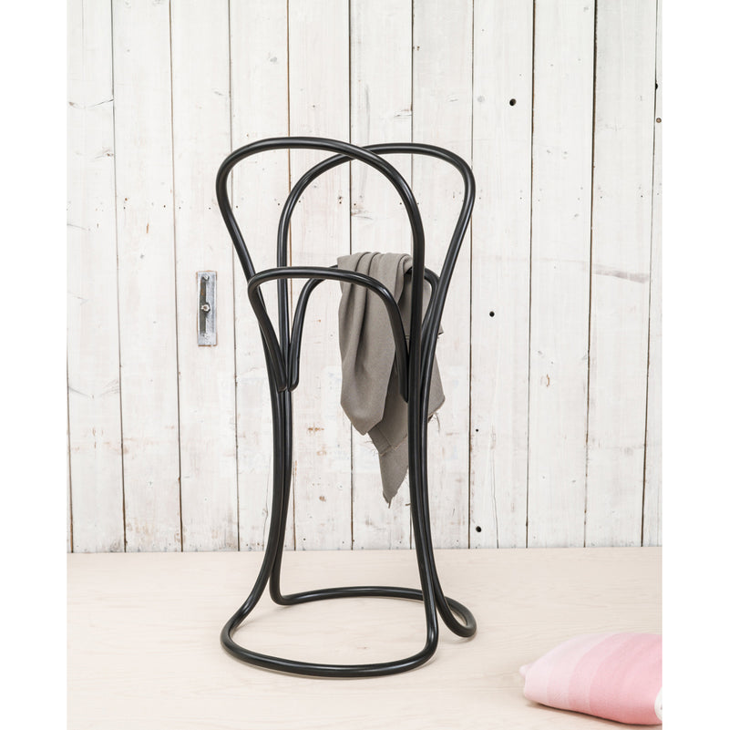 The Petalo by TON is a sleek black metal valet stand with a minimalist curved design, holding a gray garment and resting on a light wooden floor against rustic white planks. A folded pink fabric peeks out below.
