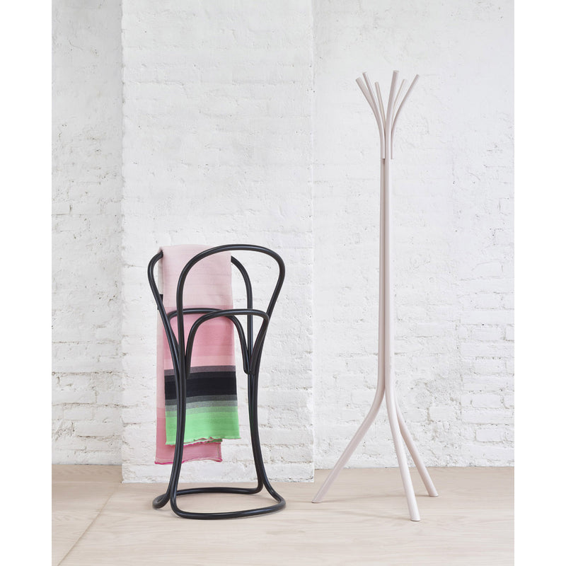 The Petalo, a black bentwood chair by TON, cradles a striped blanket in pink, black, and green. Nearby, a minimalist white coat rack with branching hooks enhances the scene against a white brick wall and light wood floor.