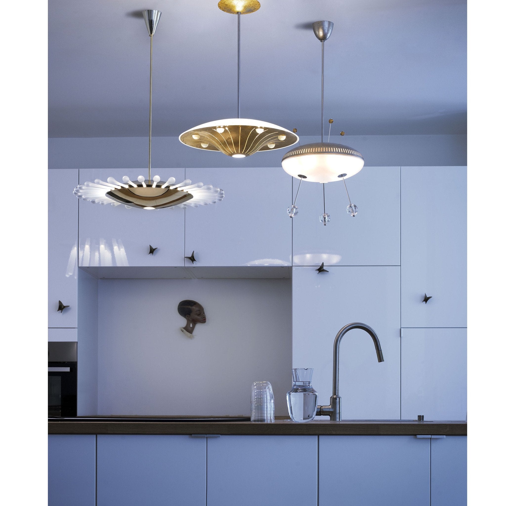 A modern kitchen showcases white cabinets, a silver faucet, and three UFO series pendant lights. A small portrait adorns the wall, while Lobmeyr glasses and a Space Ship Ellen water jug on the countertop evoke a space travel ambiance.