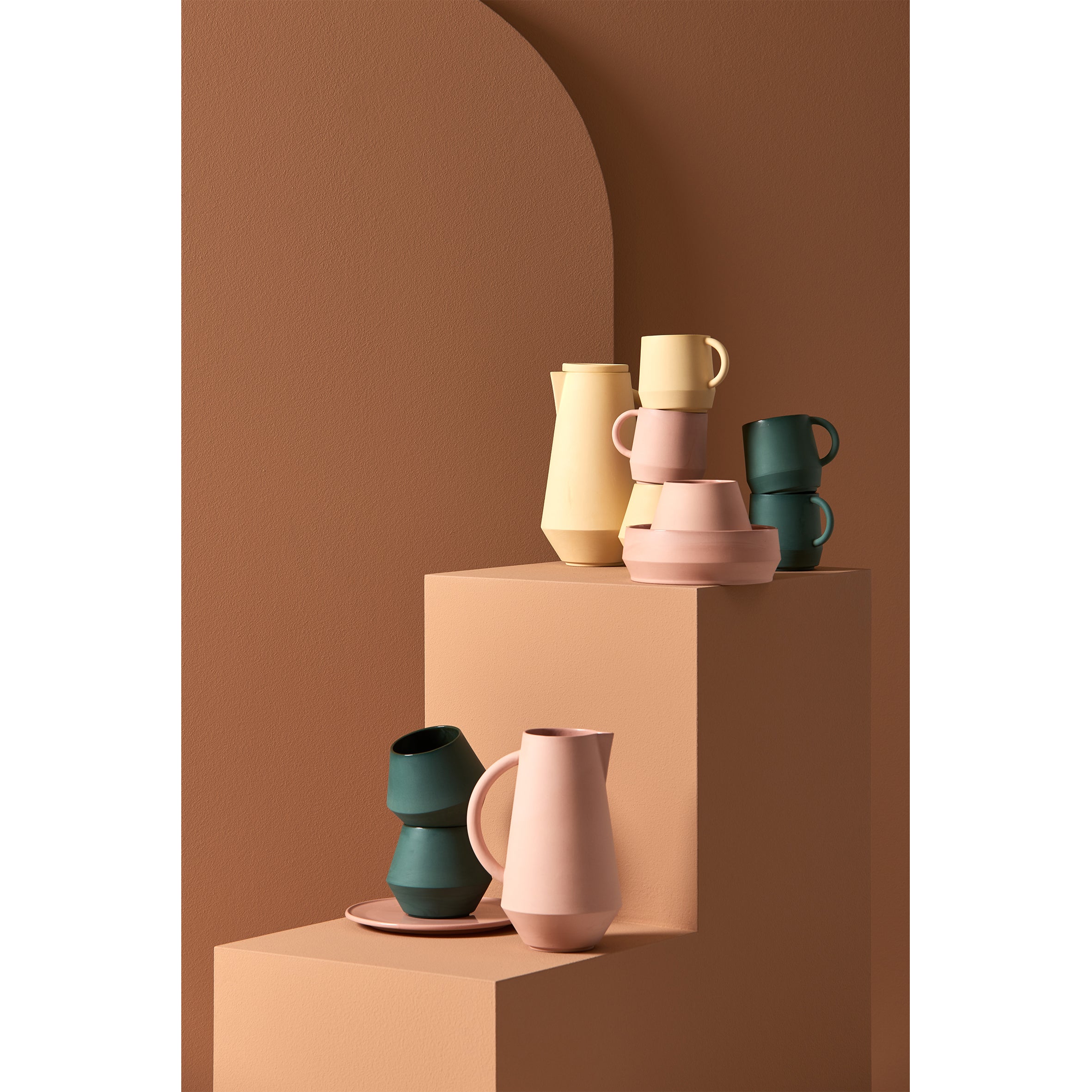 The Unison Cup by Schneid is part of a minimalist ceramic tableware collection with vases and cups in beige, green, and soft pink on geometric steps against an earthy backdrop. Featuring hand-mixed pigments, these vessels offer a modern matte finish and simple, elegant shapes.