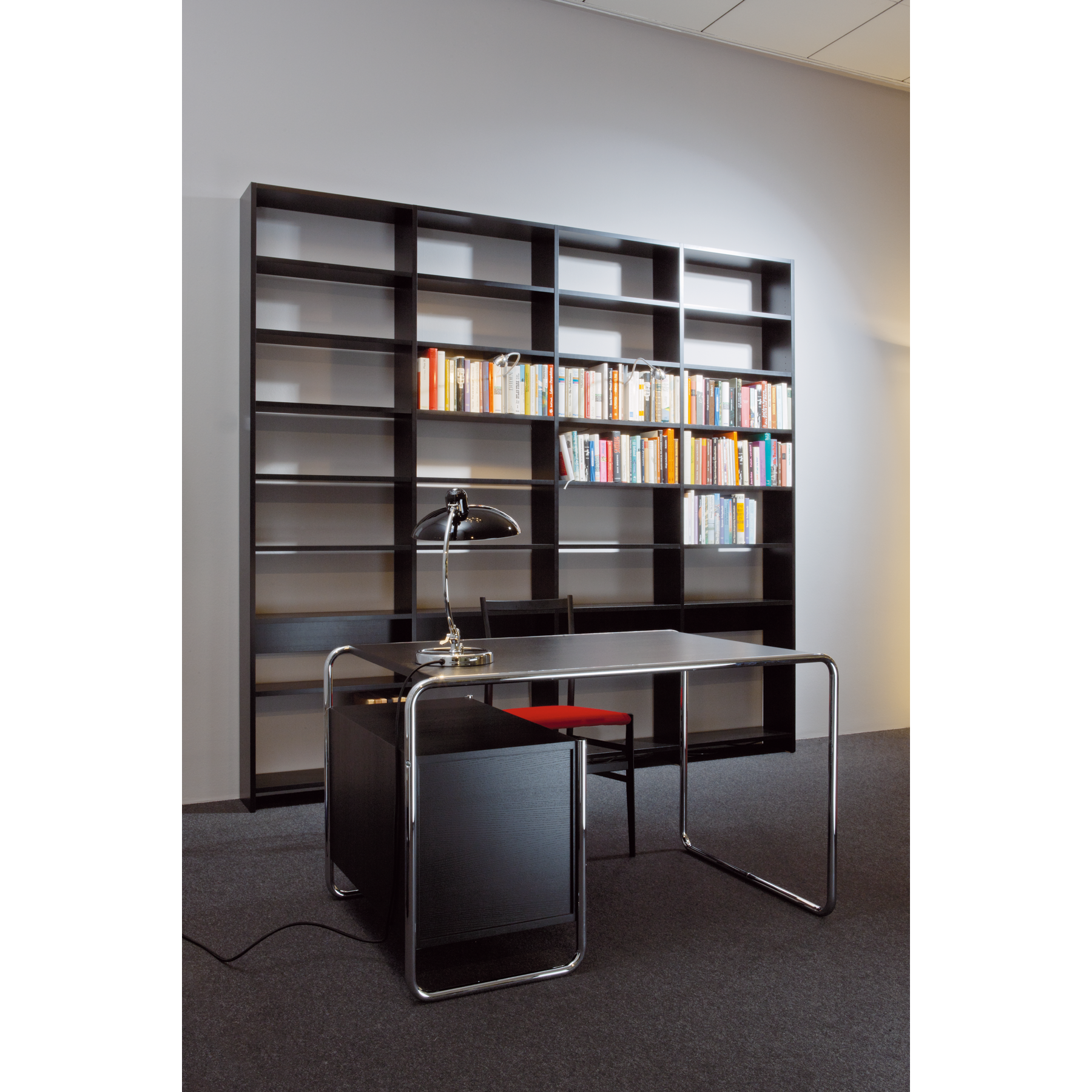 A modern office space showcases an empty black desk with metal legs paired with a black chair. A sleek desk lamp adorns the desk. In the background, the UPW Shelf by WB Form stands tall, crafted from sustainable materials and partially filled with books against a gray wall.