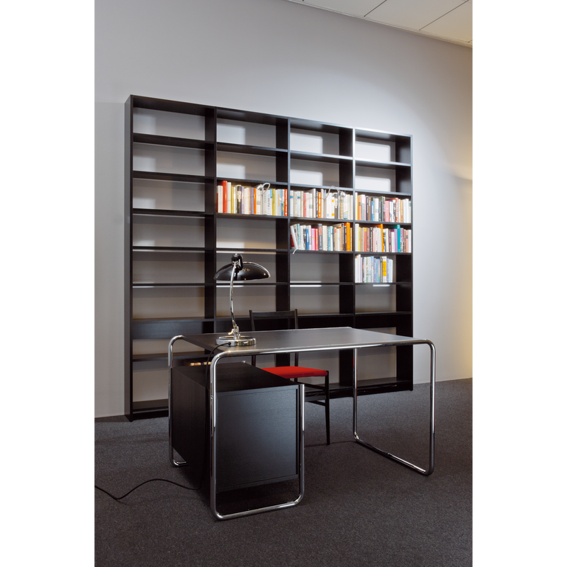 A modern office space showcases an empty black desk with metal legs paired with a black chair. A sleek desk lamp adorns the desk. In the background, the UPW Shelf by WB Form stands tall, crafted from sustainable materials and partially filled with books against a gray wall.