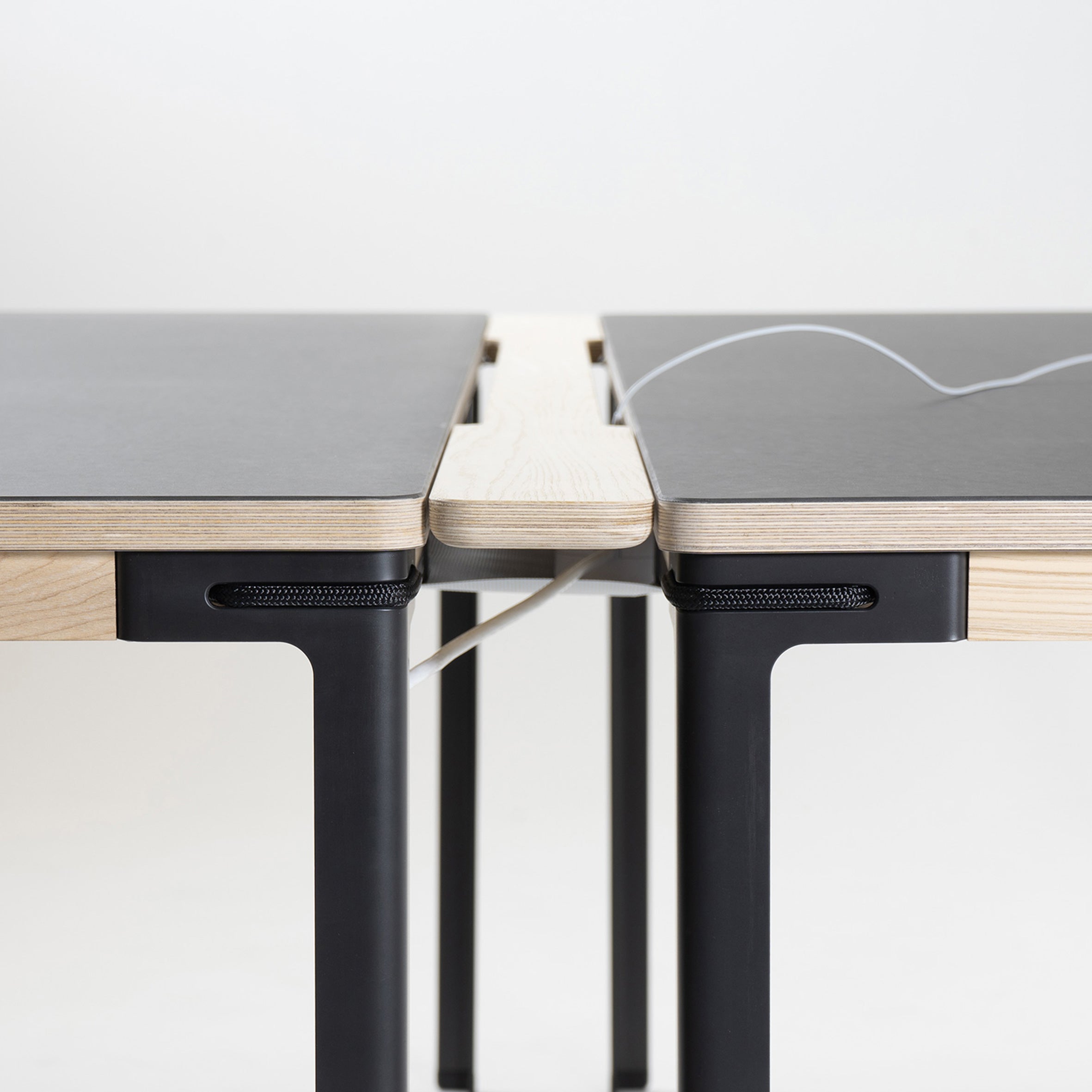 A close-up shows two modular Moormann tables featuring light wood edges and black surfaces, joined by a central wooden panel. The black table legs highlight Moormanns clever Table Connector, with a partially visible cable passage between the surfaces.