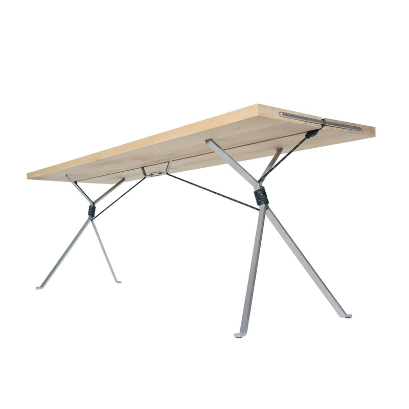The Kampenwand Table by Moormann, designed by Nils Holger Moormann, boasts a light wooden top and distinctive metal legs with X-shaped supports connected by black cords, giving it an industrial and minimalist style similar to a sleek bench.