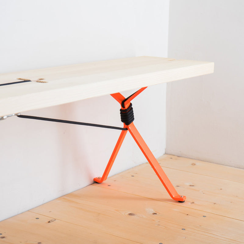 The Kampenwand Table by Moormann features bright orange metal legs, black support cords, and a minimalist wooden design. Resting on a light wooden floor against a white wall, it embodies a modern and airy aesthetic reminiscent of Nils Holger Moormanns style.