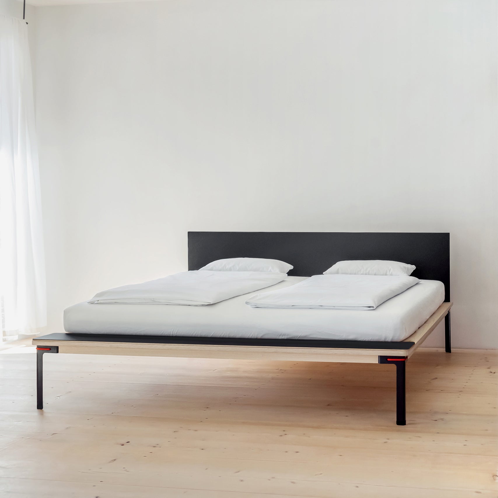 A minimalist bedroom showcases the Moormann Seiltänzer Bed with delicate steel legs, dressed in white bedding and tidy pillows. The light wood flooring and softly illuminated white walls accentuate the clean, modern aesthetic.