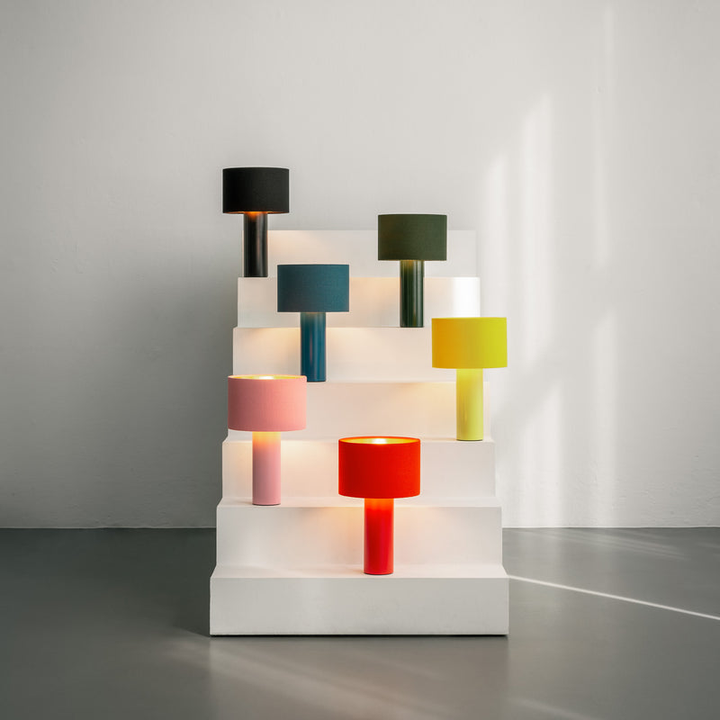 A minimalist display showcases Victor Foxtrots ALL ROUND MINI table lamps with vibrant wool fabric lampshades, neatly arranged on white stepped shelves. Each lamp, featuring a cylindrical base and colorful matching shade, provides a striking contrast against the neutral backdrop.