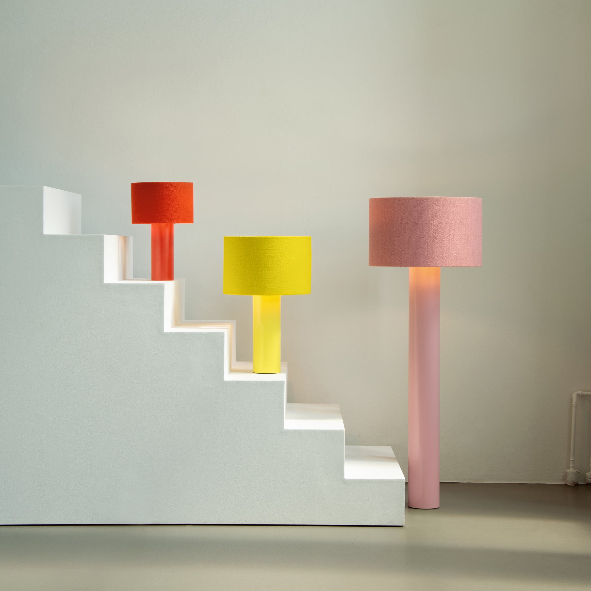 Three ALL ROUND Table Lamps by Victor Foxtrot in orange, yellow, and pink stand on a stair-like white platform against a plain wall. Each lamp has a wool fabric shade and is illuminated to create a warm, colorful ambiance.