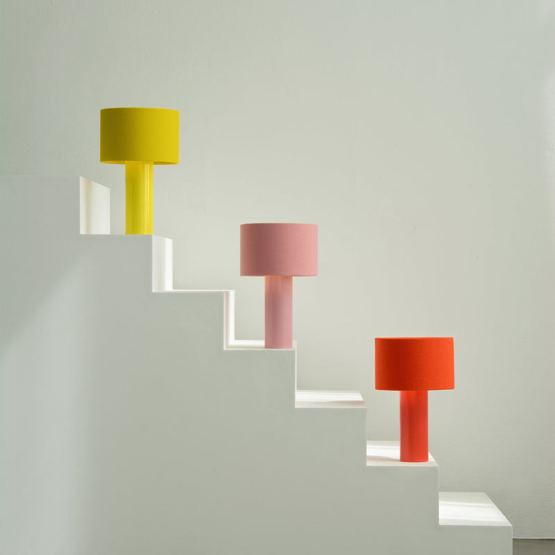 Three vibrant table lamps, including the ALL ROUND MINI by Victor Foxtrot, rest on ascending white steps against a light green wall. Their wool fabric lampshades—in yellow, pink, and orange—complement the sleek cylindrical bases perfectly.