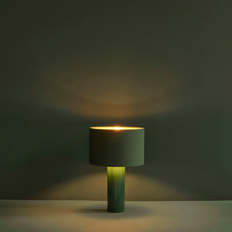 The ALL ROUND Table Lamp by Victor Foxtrot, with a dark green wool fabric shade and powder-coated steel base, emits a warm glow. Positioned on a smooth surface against a matching wall, it creates a minimalist and serene ambiance with gentle shadows.