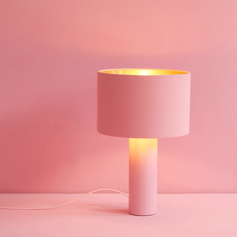 The ALL ROUND Table Lamp by Victor Foxtrot features a pink powder-coated steel base and a wool fabric lampshade, casting a warm glow. Placed on a matching pink background, it achieves a cohesive monochromatic look, with the cord visible at the base.