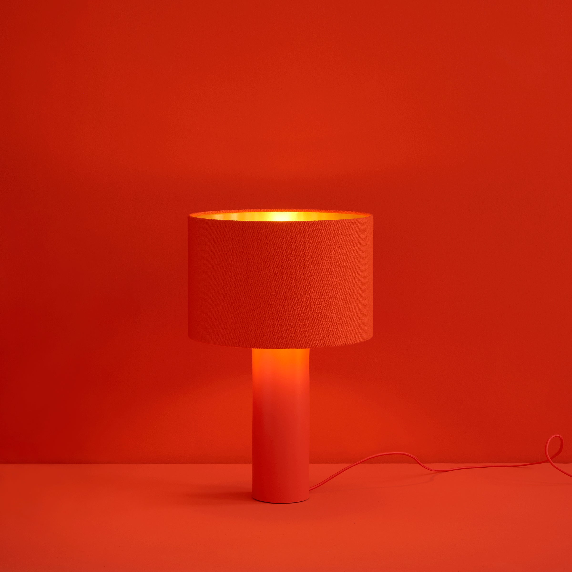 The ALL ROUND Table Lamp by Victor Foxtrot features a red cylindrical wool lampshade and a powder-coated steel base. It emits a warm glow against a matching red background and includes a thin red power cord.