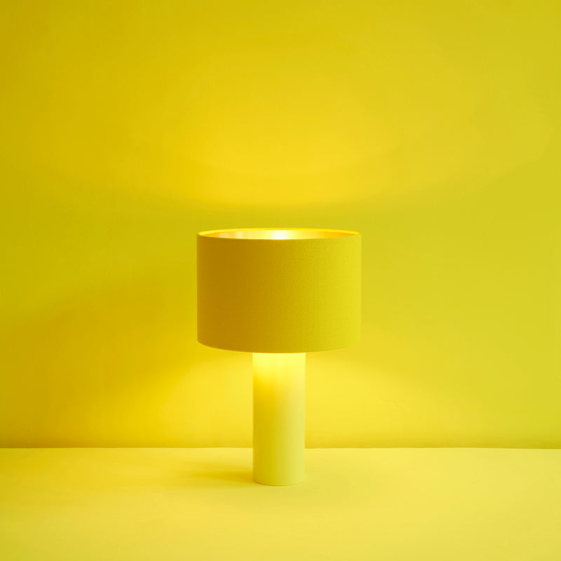 The ALL ROUND Table Lamp by Victor Foxtrot showcases a wool fabric lampshade, resting on a cylindrical, powder-coated steel base. With its minimalist yellow design and soft glow, it radiates warmth against a matching backdrop.