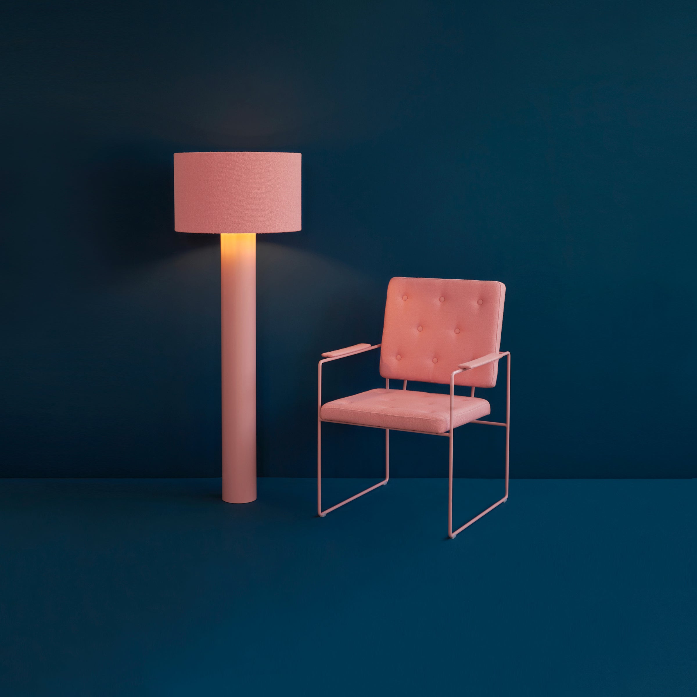 A minimalist room showcases a pink padded chair with thin metal arms and legs, upholstered in soft wool. Next to it stands the ALL ROUND Floor Lamp by Victor Foxtrot, casting warm light. The deep teal background and floor create a striking color contrast.