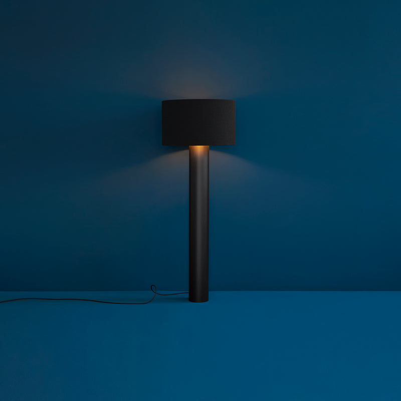 The ALL ROUND Floor Lamp by Victor Foxtrot stands against a dark blue wall, featuring a cylindrical base and round shade that cast a warm light. Resembling cozy wool fabric, it offers subtle illumination with an elegant black cord extending from the base.