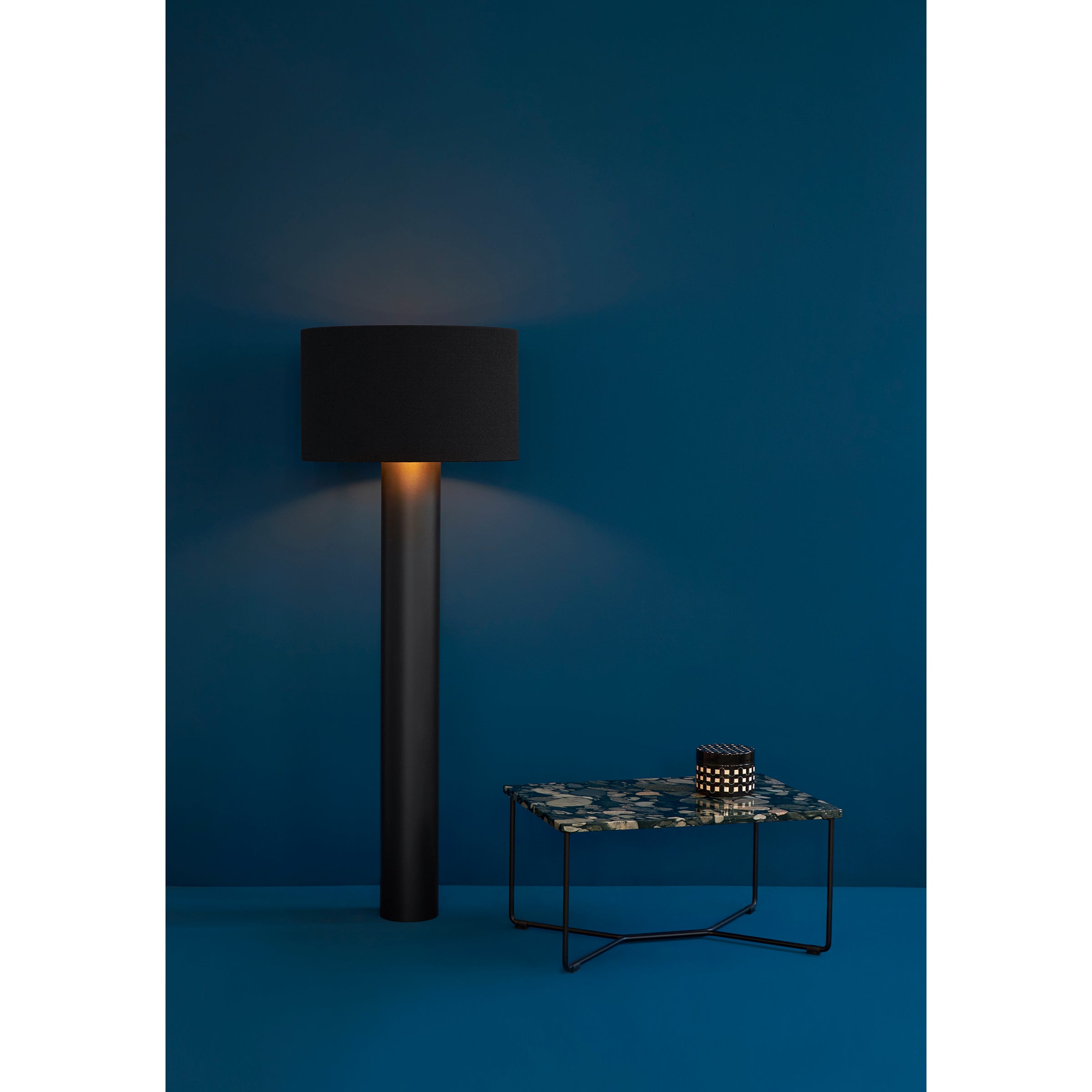 A minimalist room features a Victor Foxtrot ALL ROUND black floor lamp emitting warm light on a blue wall. Beside it, a small floral-patterned table holds an abstract decor piece. The blue wool flooring complements the wall, creating a cozy atmosphere.