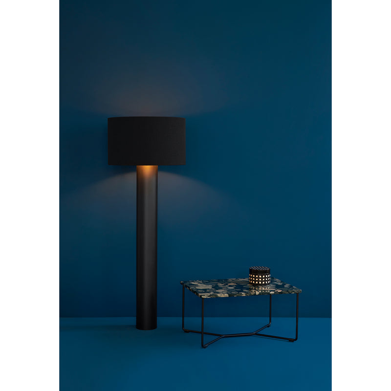 A minimalist room features a Victor Foxtrot ALL ROUND black floor lamp emitting warm light on a blue wall. Beside it, a small floral-patterned table holds an abstract decor piece. The blue wool flooring complements the wall, creating a cozy atmosphere.