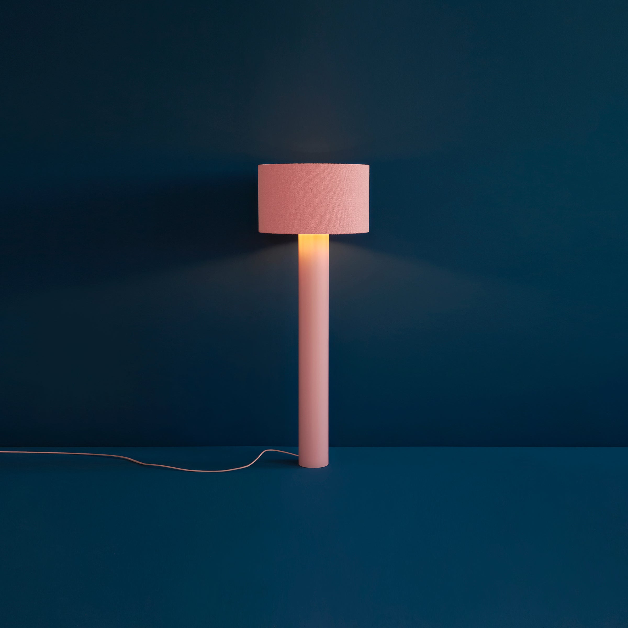 The ALL ROUND Floor Lamp by Victor Foxtrot features a pink cylindrical base and matching wool-wrapped lampshade, casting a warm glow against a deep blue wall. A white power cord extends from the base.