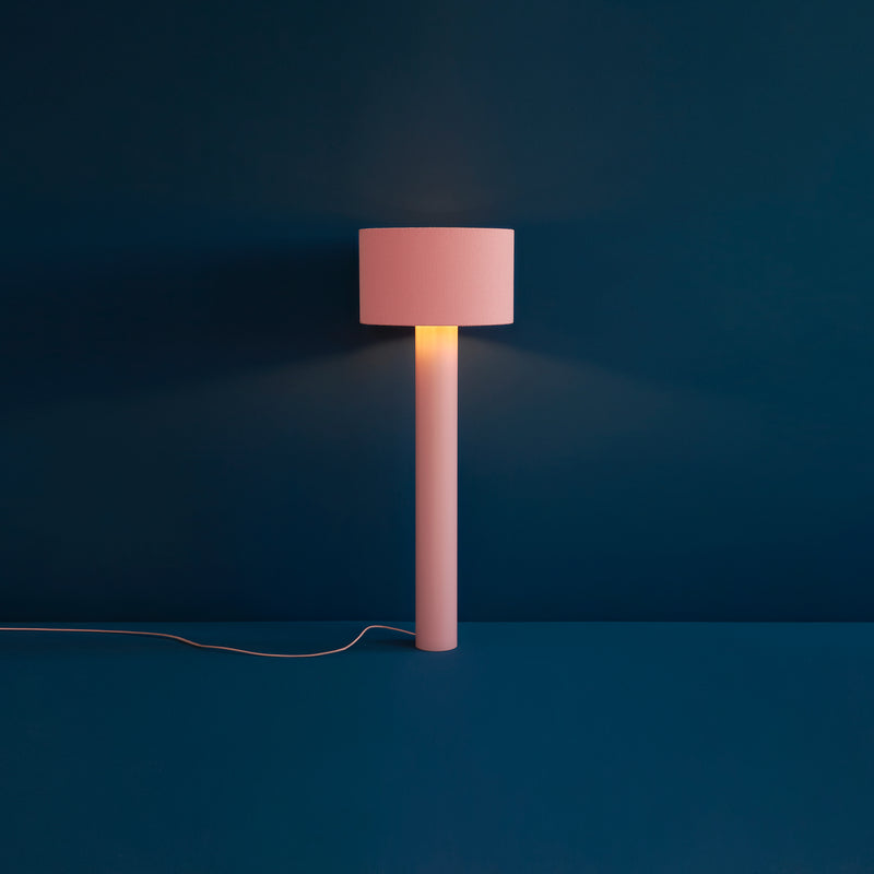 The ALL ROUND Floor Lamp by Victor Foxtrot features a pink cylindrical base and matching wool-wrapped lampshade, casting a warm glow against a deep blue wall. A white power cord extends from the base.