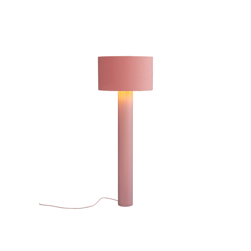 The ALL ROUND Floor Lamp by Victor Foxtrot is a pink lamp with a cylindrical base and wool fabric drum shade, emitting warm light. A power cord extends from the base, all set against a plain white background.