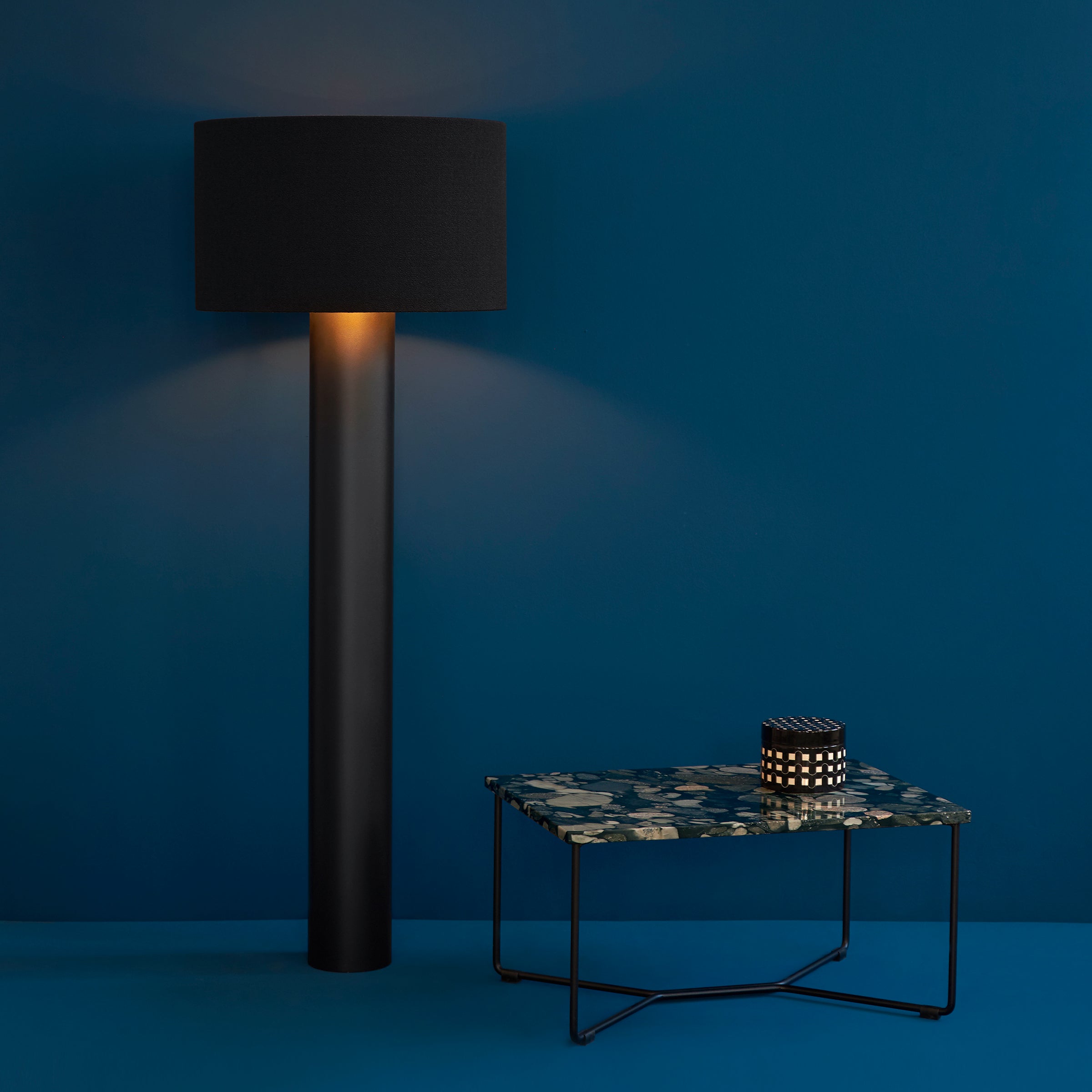 A sleek black floor lamp emits a warm glow next to a Nero Marinace-themed POI SINGLE MARBLE Side Table from Victor Foxtrot, featuring a striking blue and black patterned top. A small decorative item sits on the table, all against a deep blue wall and floor backdrop.