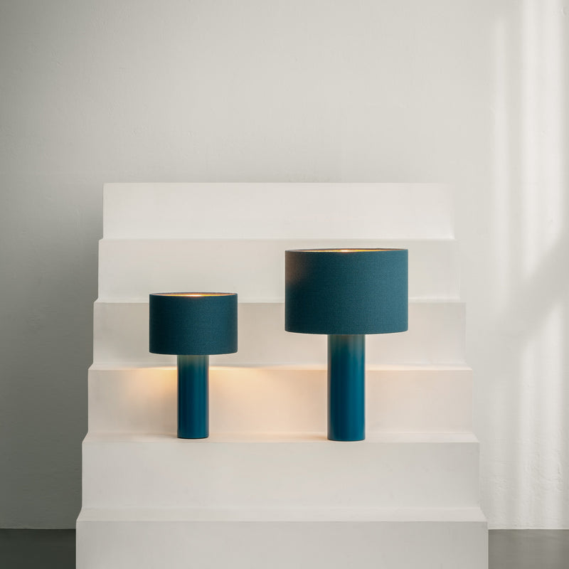 Two ALL ROUND MINI Table Lamps by Victor Foxtrot, with wool fabric lampshades, rest on white stepped pedestals against a light gray wall. They feature cylindrical bases and drum-shaped shades, casting a soft glow around them.