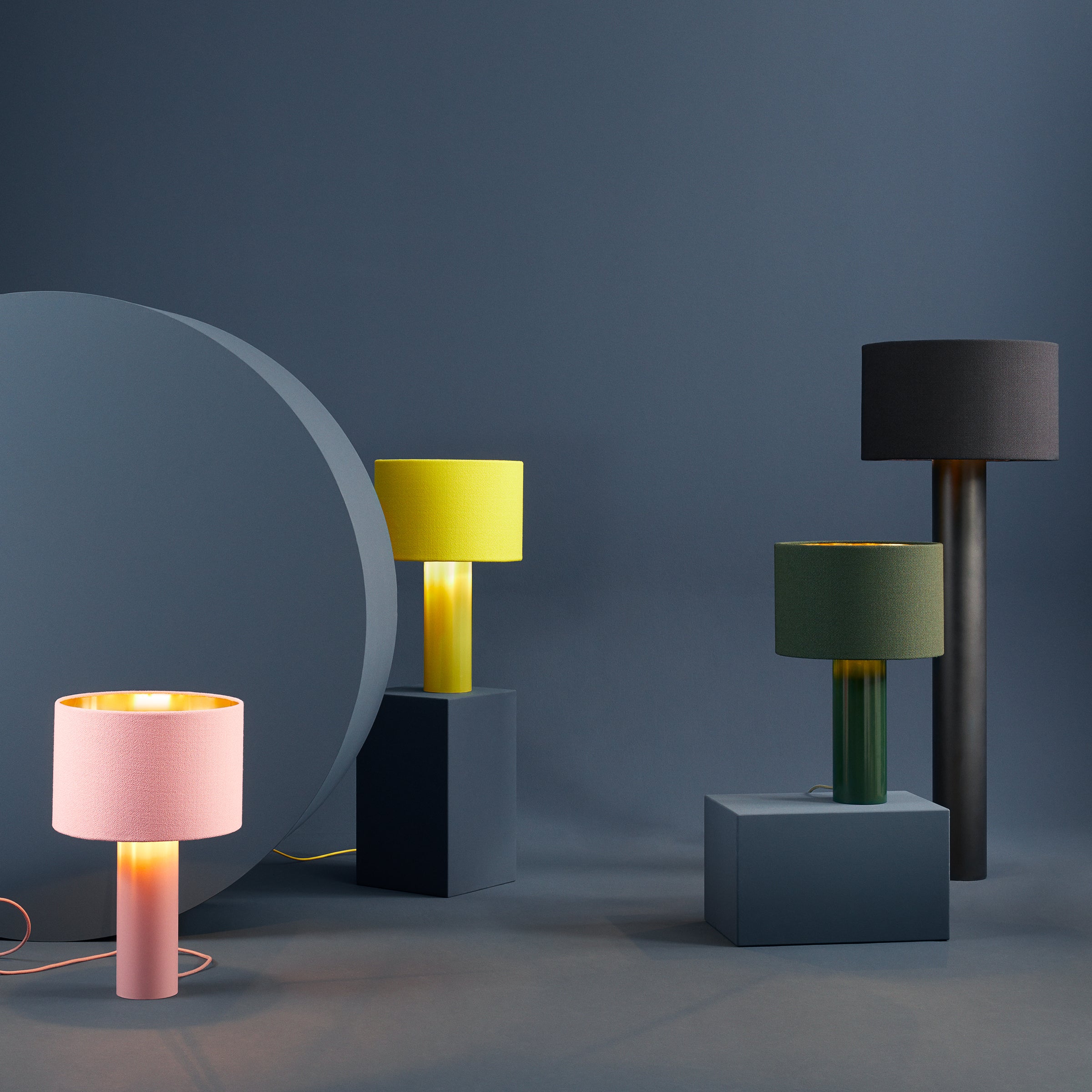 Four ALL ROUND Floor Lamps by Victor Foxtrot, featuring cylindrical bases and colorful shades in pink, yellow, green, and black, are artfully arranged on dark geometric pedestals against a soft blue background, casting a warm light that enhances their vibrant hues.