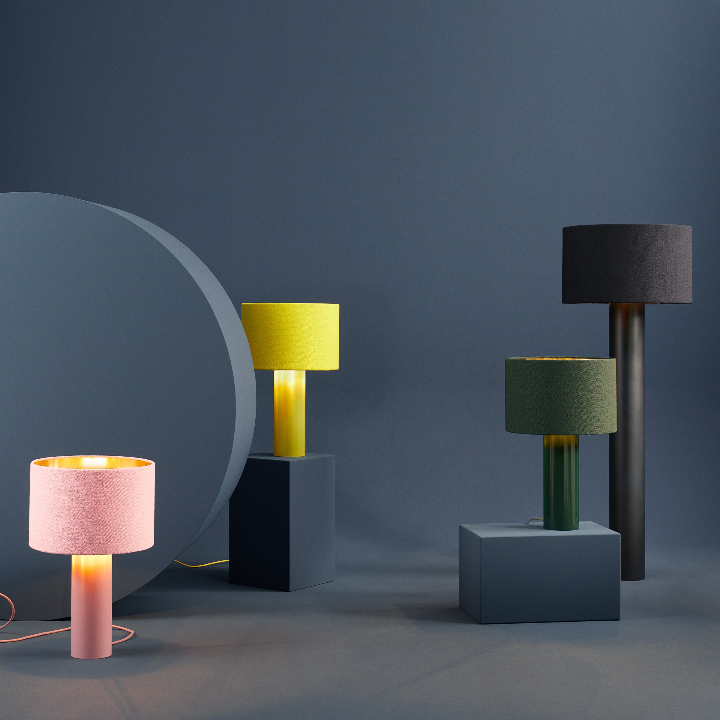 Four ALL ROUND Table Lamps by Victor Foxtrot feature cylindrical powder-coated steel bases and wool fabric shades in pink, yellow, green, and black. Arranged on geometric platforms against a dark blue backdrop, they create a modern artistic display with warm lighting.