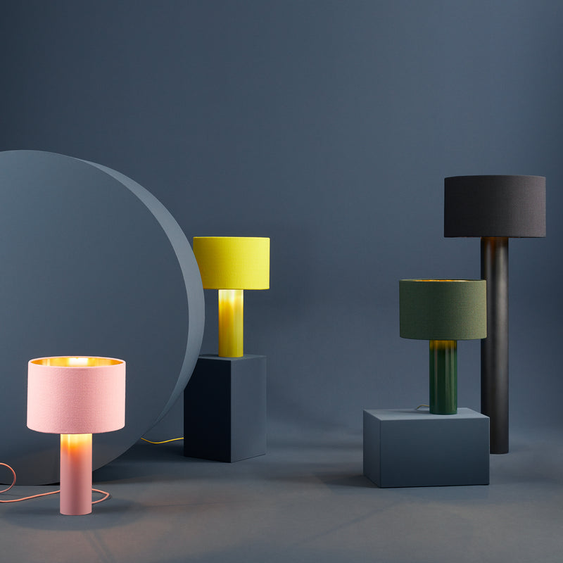 Four ALL ROUND Floor Lamps by Victor Foxtrot, featuring cylindrical bases and colorful shades in pink, yellow, green, and black, are artfully arranged on dark geometric pedestals against a soft blue background, casting a warm light that enhances their vibrant hues.