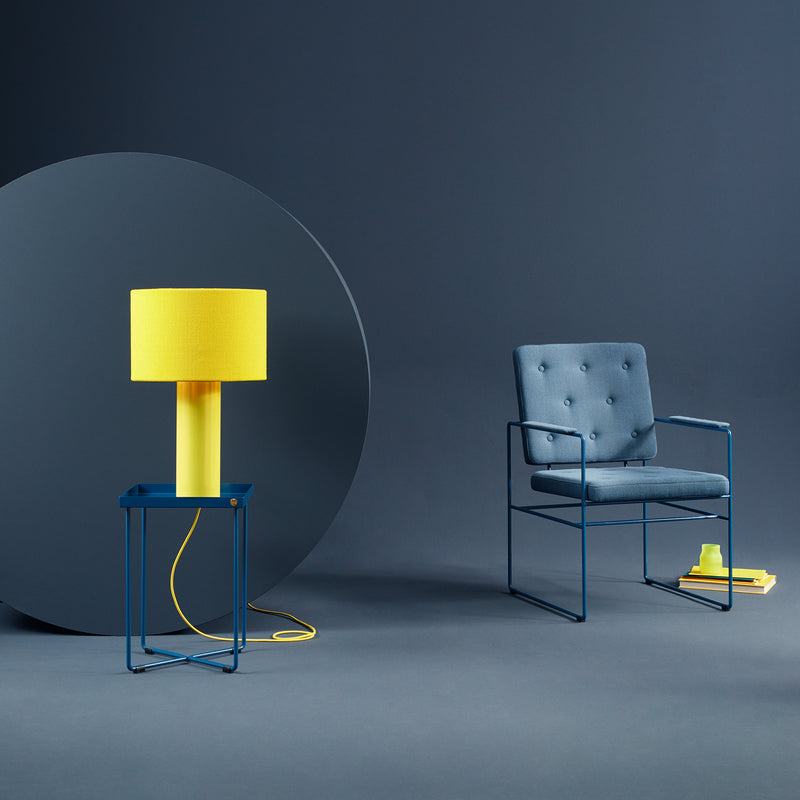 A modern interior features a blue cushioned armchair with yellow books and a cup beside it. Nearby, the POI MINI Side Table by Victor Foxtrot holds a lamp with a vibrant yellow shade, set against a dark blue circular backdrop.