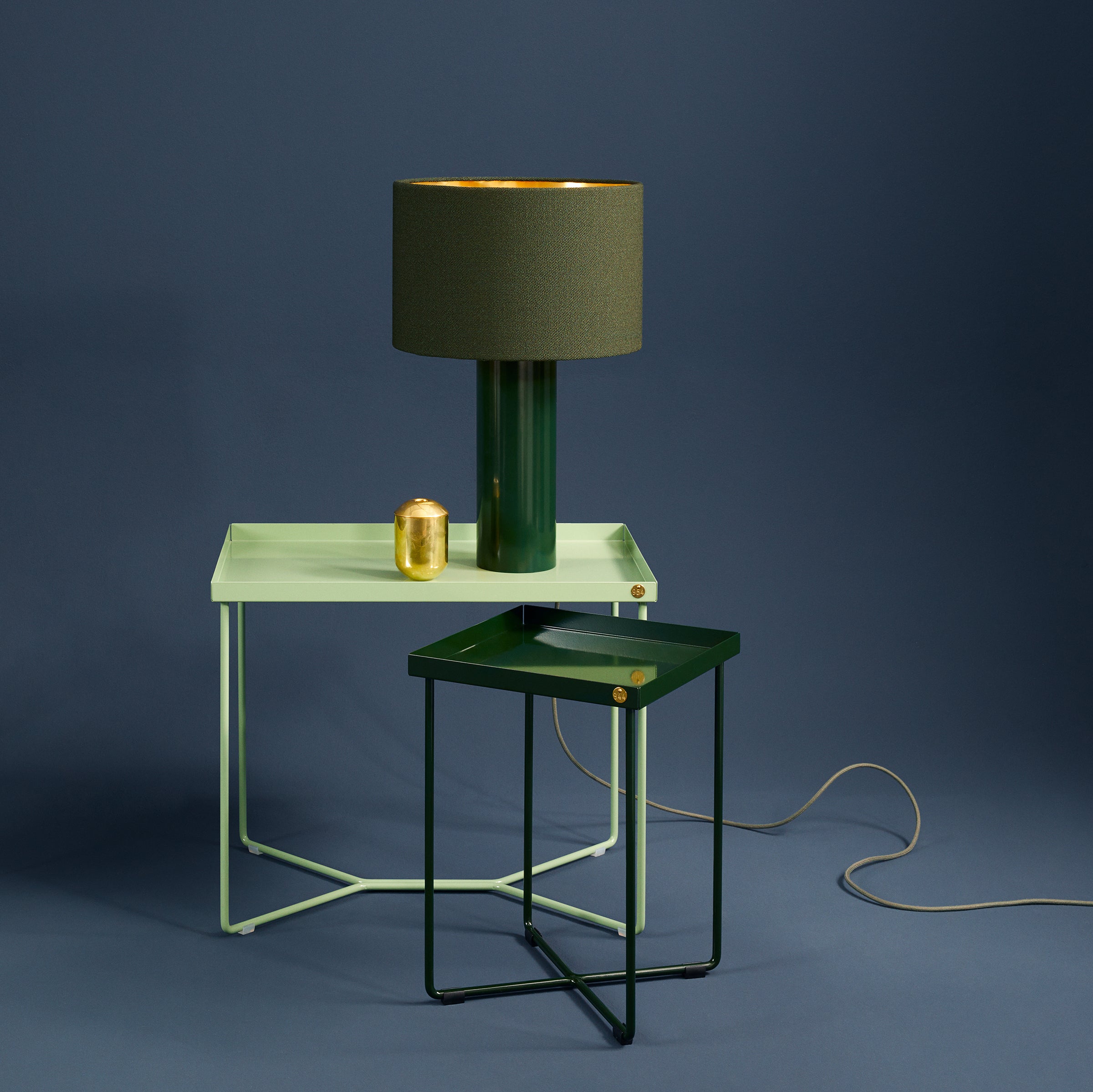 A minimalist setup features a Victor Foxtrot POI MINI mint green side table and a dark green bedside table. Atop sit a dark green lamp with a gold interior and a small gold container, with its power cord set against a smooth, muted blue backdrop.