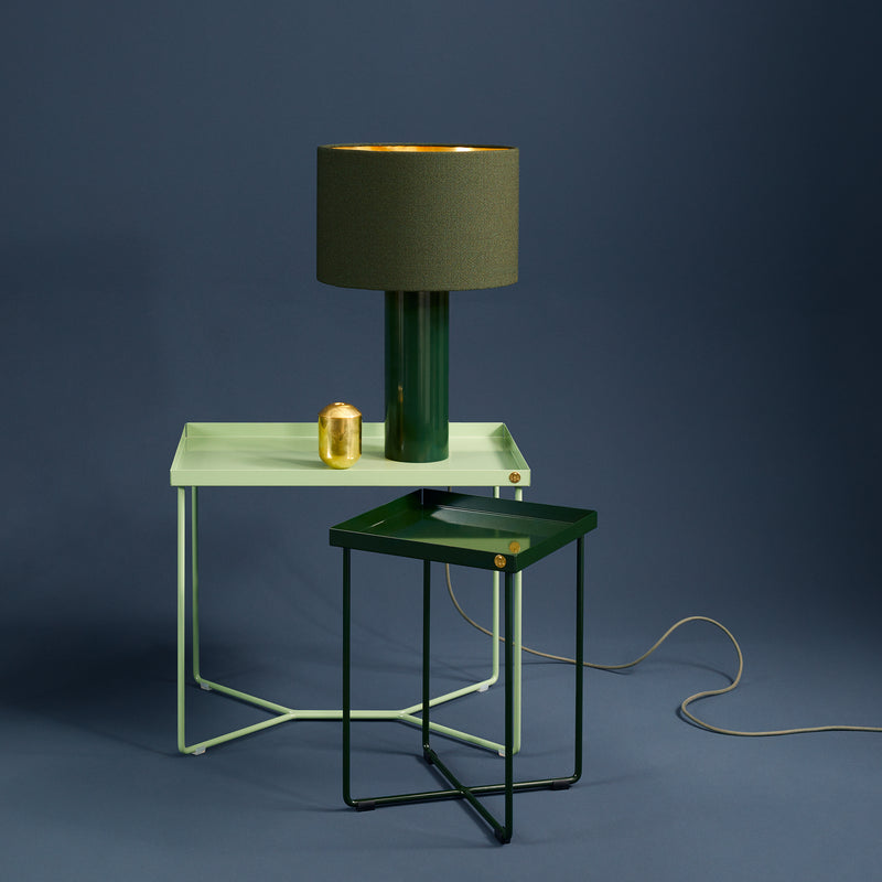 A minimalist setup features a Victor Foxtrot POI MINI mint green side table and a dark green bedside table. Atop sit a dark green lamp with a gold interior and a small gold container, with its power cord set against a smooth, muted blue backdrop.